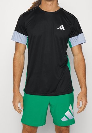 Man wearing black sports t-shirt with gray sleeves and green shorts, both featuring white Adidas logos, standing against a plain background.