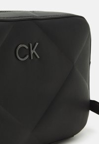 Borsa cosmetica nera trapuntata con una texture in pelle liscia. Presenta un logo grigio "CK" sul davanti e una chiusura con zip sulla parte superiore.