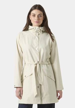 Kobieta w beżowej przeciwdeszczowej kurtce z kapturem, zapinanej na zatrzaski, z paskiem w talii i dużymi przednimi kieszeniami, stojąca na jednolitym tle.
