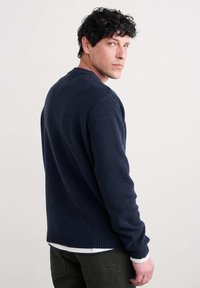 Tumši zils tekstūrēts džemperis ar apaļu kakla izgriezumu un ribotu apakšmali, vilkts virs baltas krekla, attēlots no sāna, ar redzamām roku detaļām.