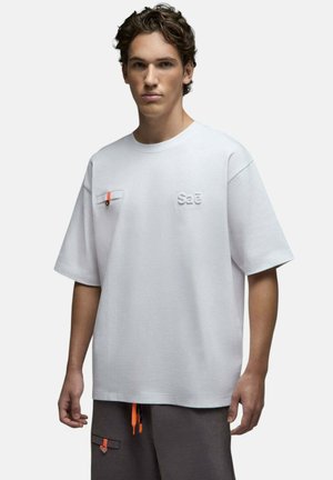 Jeune homme portant un T-shirt blanc oversize avec "Sae" embossé sur la poitrine et un pantalon foncé avec des tirettes de fermeture éclair orange et des détails de poche.