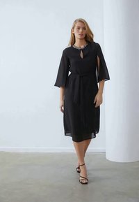 Juoda suknelė su rakto skylutės iškirpte, mėgstamais apsiaustais ir rišama juosta. Papuošta brangakmenių apykakle ir hemline žemiau kelių.
