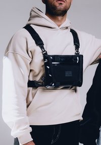 Borsa a tracolla nera con finitura lucida, cinturini regolabili, fissata con ganci metallici, indossata sopra un hoodie beige chiaro. Design minimalista.