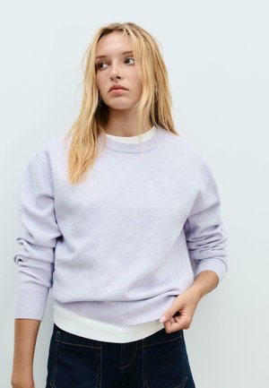 Jeune femme aux cheveux blonds, portant un pull violet clair et un jean sombre, regardant sur le côté devant un fond uni clair.