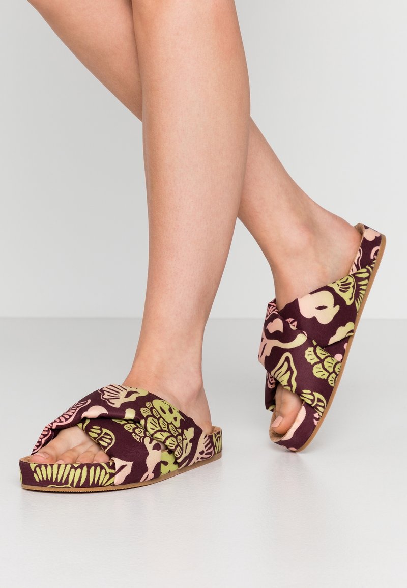 Jambes portant des sandales à enfiler avec des brides en tissu large et torsadé, présentant des motifs floraux verts et roses sur une base bordeaux.