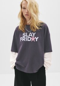 Oversized grå t-shirt med teksten "SLAY FRIDAY" og en pink stjerne. Lange hvide ærmer synlige under, afslappet pasform, lavet af bomuldsmateriale.