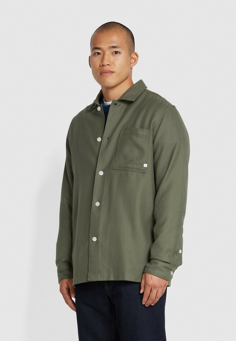 Olijfgroene button-up overhemd van gestructureerde stof, met een borstzak en witte knopen. Draag over een marineblauw shirt, met lange mouwen.