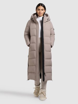 Long, light taupe puffer coat with a hood, quilted design, front zipper, black accents, and side pockets. Worn over beige outfit and boots.