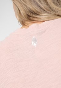 Světle růžový svetr s jemnou texturou. Na zadním výstřihu se nachází decentní květinový motiv. Kulatý výstřih a žebrované okraje.