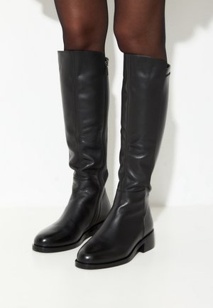 Tall black leather boots worn with sheer black tights, standing against a plain white background.