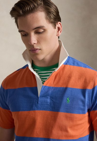 Πουκάμισο ράγκμπι με γιακά και οριζόντιες μπλε και πορτοκαλί ρίγες. Διαθέτει πράσινο κεντητό λογότυπο στο στήθος και λευκό γιακά με πουκάμισο με πράσινη ρίγα από κάτω.