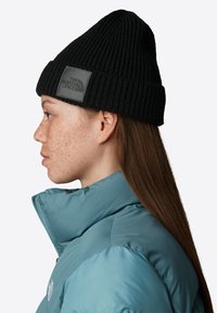 Mustat neulepipo, jossa on ribbitekstuuri ja nahkainen logo-osa, yhdistettynä teal-väriseen toppatakkiin, jossa on korkea kaulus.