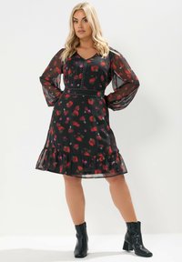 Robe noire à fleurs avec des manches longues transparentes, un ourlet froncé et des détails en perles à la taille. Présente des motifs de fleurs rouges et roses.