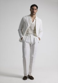 Traje de lino blanco que incluye un blazer ajustado, pantalones plisados y una camisa polo de punto color beige, complementado con mocasines oscuros.