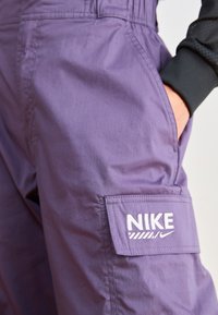 Purple cargo pants with a white Nike logo on the pocket, worn with a black long-sleeve shirt, hand resting inside the pocket.