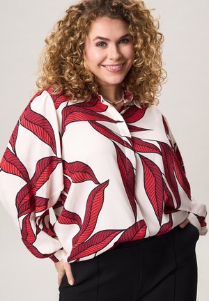 Lachende vrouw met krullend blond haar, gekleed in een wit hemd met grote rode bladpatronen en zwarte broek, handen in de zakken.