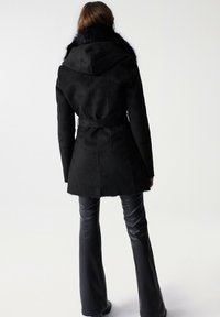 Mujer vistiendo un abrigo negro con cinturón y cuello de piel, y pantalones de cuero negros acampanados, de pie de espaldas frente a un fondo sencillo.