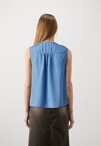 Vaaleansininen ilman hihoja oleva paita, joka on valmistettu pehmeästä kankaasta. Paita on varustettu kerätyllä selkädesignilla ja pyöreällä kaula-aukolla. Yhdistetty tummanruskeaan hameeseen.