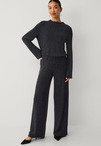 Zwart gebreide top met patroon en lange mouwen, gecombineerd met broek met wijde pijpen. Beide kledingstukken hebben een subtiel gespikkeld ontwerp in wit. Zwarte puntige schoenen.