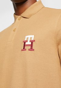 Brązowa koszulka polo z fakturowanej tkaniny, z wyszywanym białym i burgundowym logo składającym się z trzech liter na piersi.