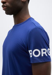 Άνδρας που φορά μπλε κοντομάνικο πουκάμισο με λευκά διάτρητα γράμματα "BORG" στο μανίκι, εμφανιζόμενος από τον ώμο μέχρι τον λαιμό.