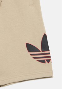 Calções bege feitos de mistura de algodão com textura macia. Apresentam um grande logótipo preto com detalhes vermelhos na perna esquerda.