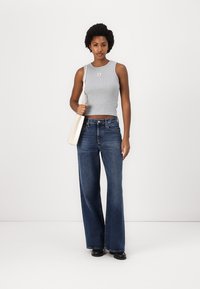 Šedý bezrukávový crop top s logem, spárovaný s volnými tmavomodrými džíny. Modelka drží krémovou crossbody tašku. Hladká textura tkaniny, neformální design.