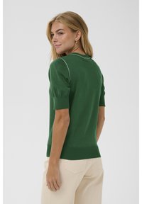 Žena s blond vlasy, oblečená v zeleném svetru s krátkým rukávem a béžových kalhotách, stojí bokem před jednotvárným světlým pozadím.