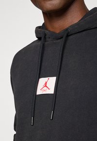 Zwarte hoodie met een gestructureerde afwerking, voorzien van een rode Jordan-logo patch en zwarte trekkoorden met plastic uiteinden.