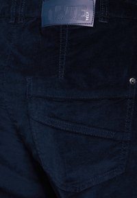 Marine corduroy broek met een getextureerde stof, achterzak met stiksel detail, en een label aan de tailleband met reliëftekst.