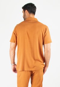 Chemise à manches courtes en tissu orange brûlé, présentant une texture lisse et une coupe décontractée, affichée de dos sur fond blanc.