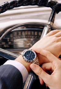 Rostamattainen kronografikello, jossa tummansininen kellotaulu, hopeiset yksityiskohdat ja linkkiketjuranneke. Käsi säätämässä aikaa ranteen päällä lepäävänä.