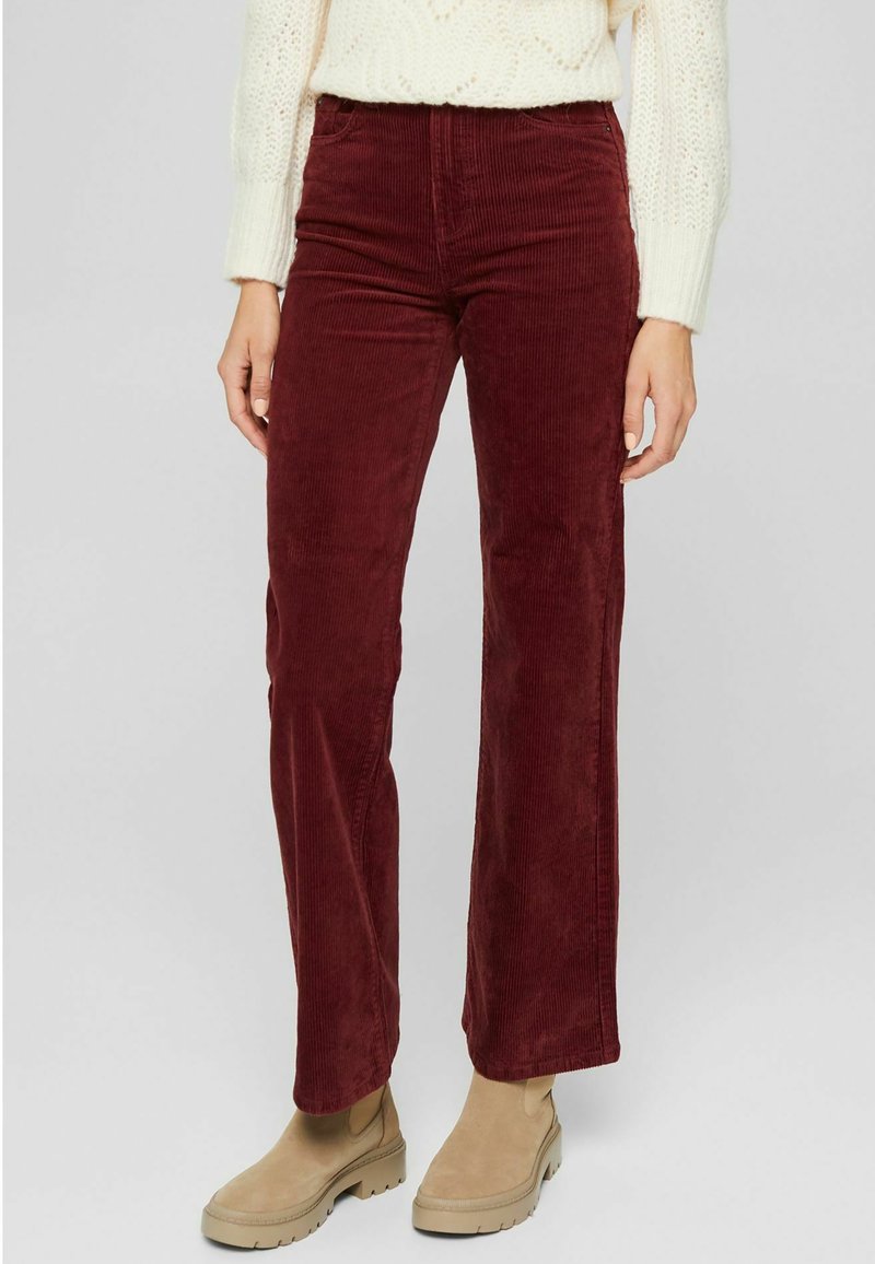 Femme portant un pantalon en velours côtelé bordeaux, taille haute et jambes droites, avec des bottines beige à la cheville et un pull en maille blanc.
