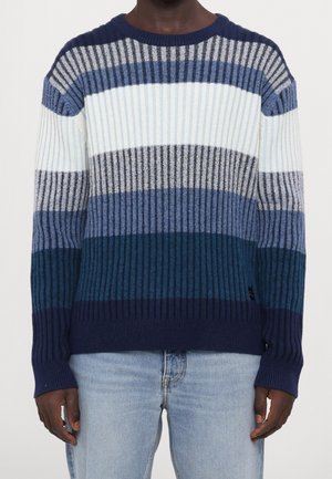 Mann trägt einen gerippten Strickpullover mit horizontalen Streifen in Blau-, Grau- und Weißtönen, kombiniert mit hellblauen Jeans.