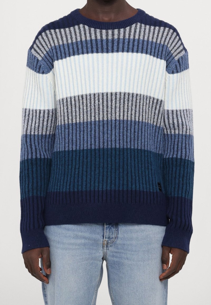 Homme portant un pull en maille côtelée à rayures horizontales dans des tons de bleu, gris et blanc, associé à un jean bleu clair.