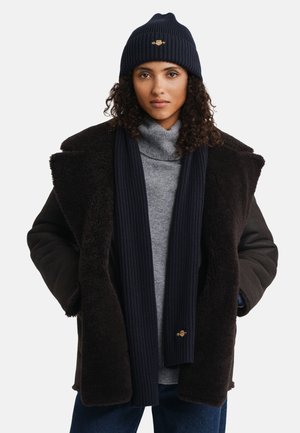 Dunkelbrauner Lammfellmantel mit einem schwarzen Pullover, marineblauem, geripptem Schal und passender Mütze. Die Mütze hat ein Logodetail auf der Vorderseite.