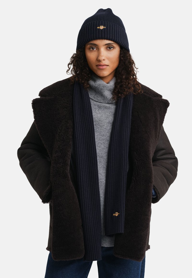 Donkerbruine schapenvachtjas met een zwarte trui, een marineblauwe geribbelde sjaal en een bijpassende muts. De muts heeft een logo op de voorkant.