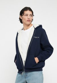 Sweat à zip marine avec une doublure en fausse fourrure blanche, poches avant et nom de la marque brodé. Comprend une capuche et des poignets côtelés.