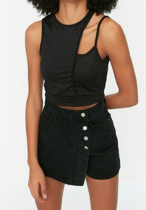 Frau trägt ein schwarzes asymmetrisches ärmelloses Crop-Top und einen hoch taillierten schwarzen Jeansrock mit silbernen Knöpfen vorne.