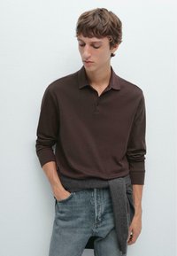 Bruine langemouw polo met een drieknops sluiting, gecombineerd met lichtblauwe jeans. Een donkergrijze trui is om de taille gebonden.