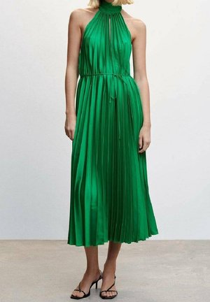 Grünes, plissiertes, ärmelloses Kleid mit hohem Kragen und Kordelzug in der Taille, aus glattem Stoff. Knöchellang, kombiniert mit schwarzen Sandalen mit dünnen Riemen.