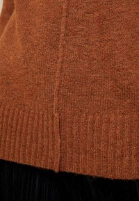 Maglione a maglia color ruggine con orlo a coste, che mostra un tessuto strutturato e un dettaglio di cucitura laterale. Il tessuto ha un aspetto morbido e peloso.