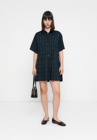 Kurzärmliges kariertes Kleid in Dunkelgrün und Marineblau mit einer durchgehenden Knopfleiste und einem ausgestellten Rock. Getragen mit burgunderfarbenen Ballerinas und einer schwarzen Tasche.