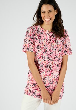 Donna sorridente che indossa una camicetta floreale rosa a maniche corte e pantaloni bianchi, in piedi di fronte a uno sfondo grigio chiaro.