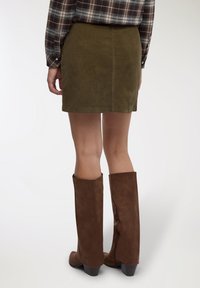 Minigonna in velluto a coste verde oliva con una texture liscia. Abbinata a stivali marroni al ginocchio con un tacco sottile e una cerniera laterale.
