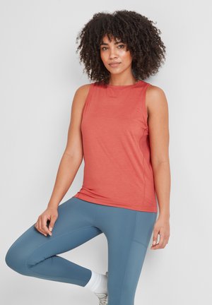 Woman with curly hair wearing a sleeveless red top and blue leggings, standing in a balanced pose against a white background.