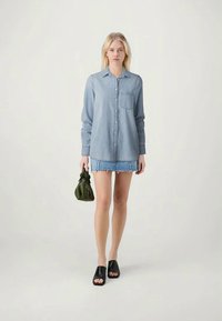 Camisa de ganga com botões e bolso no peito, combinada com uma saia de ganga azul, sandálias pretas de salto e uma pequena bolsa verde.