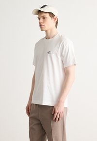 White cotton t-shirt with short sleeves and a small black logo on the left chest. Paired with beige trousers and a cream cap.