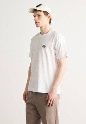 Weißes Baumwoll-T-Shirt mit kurzen Ärmeln und einem kleinen schwarzen Logo auf der linken Brust. Kombiniert mit beigen Hosen und einer cremefarbenen Kappe.