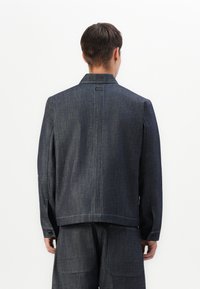 Mørk denimjakke med en simpel krave, lige snit og synlige sømme. Bagsiden har en central søm og et mærke nær halsudskæringen.